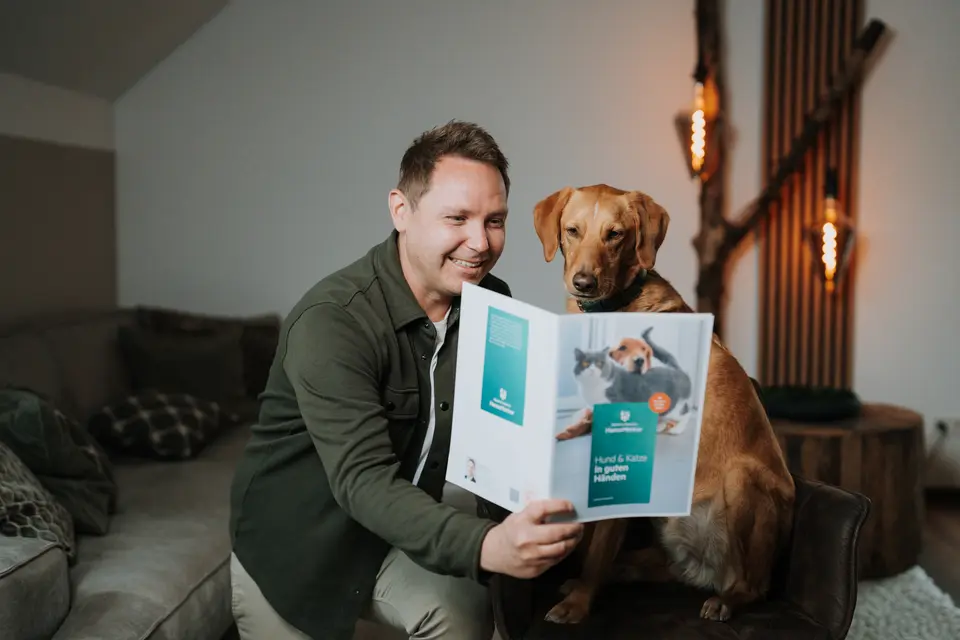 Denis Klefenz hält lachend eine HanseMerkur-Broschüre hoch, sein Hund schaut neugierig rein