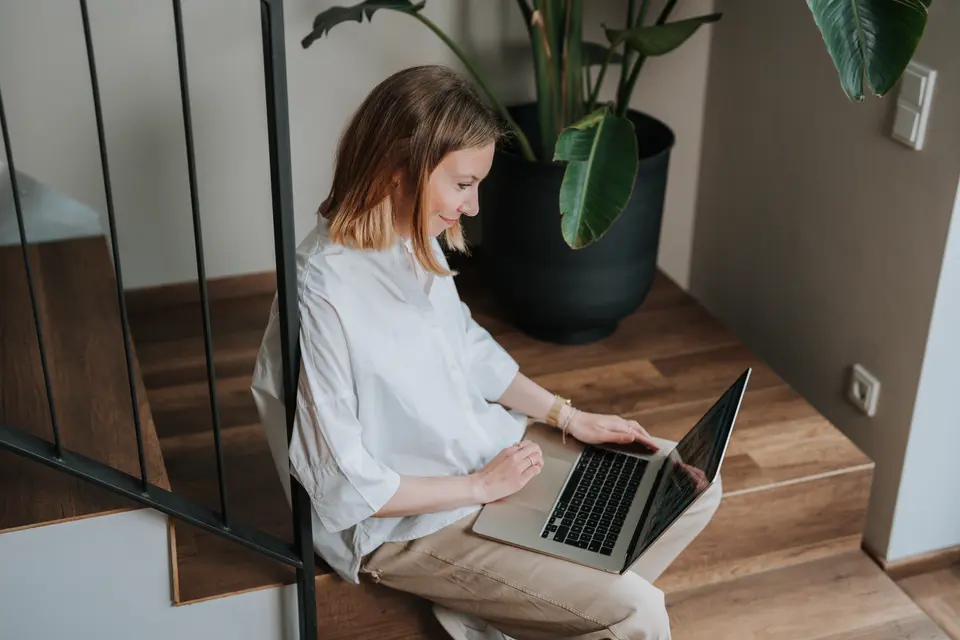 Jill sitzt auf einer Treppe und arbeitet lächelnd am Laptop neben einer großen Zimmerpflanze