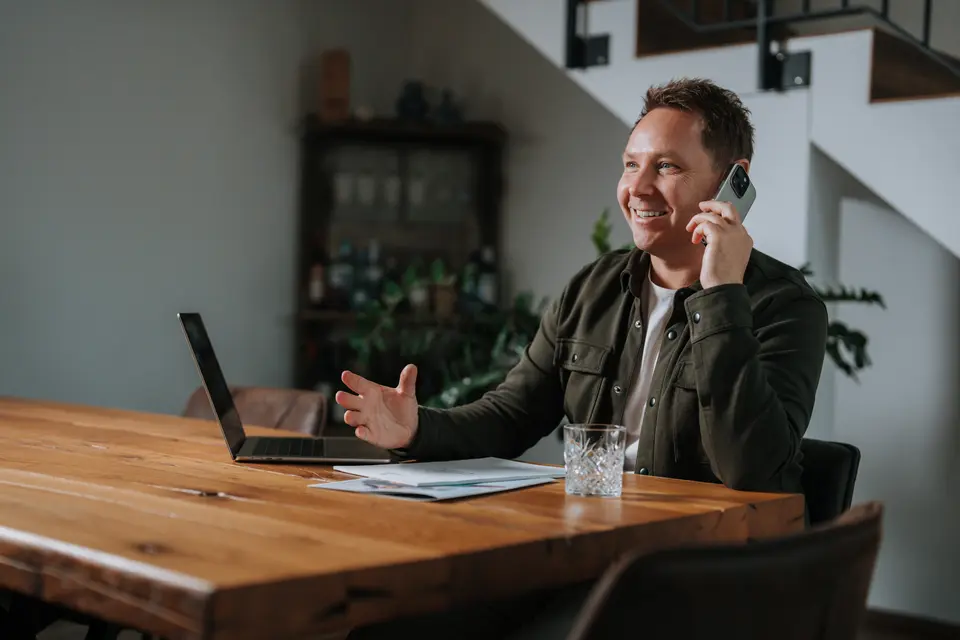 Denis Klefenz lacht am Holztisch beim Telefonieren mit Laptop und Unterlagen vor sich
