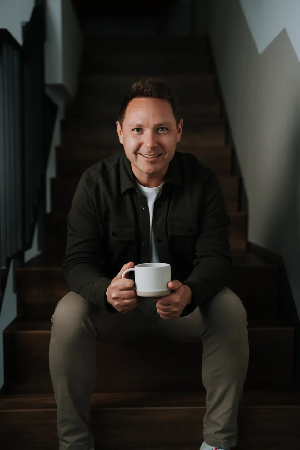 Denis Klefenz sitzt lächelnd mit einer Kaffeetasse auf einer dunklen Holztreppe