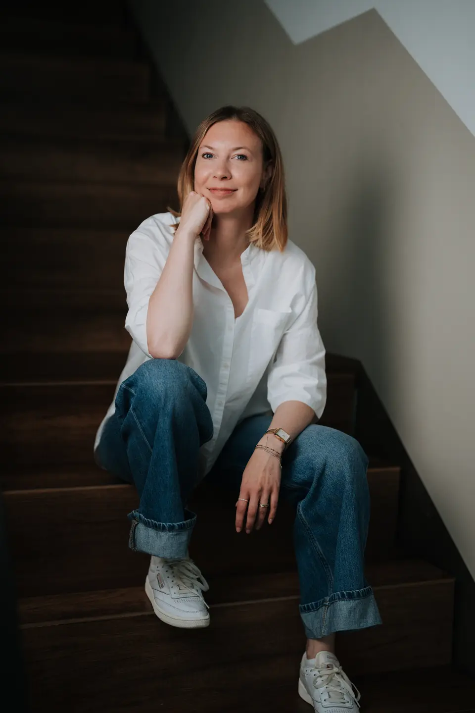 Jill, Mitarbeiterin bei Denis Klefenz, sitzt lächelnd auf einer Holztreppe in weißer Bluse und Jeans